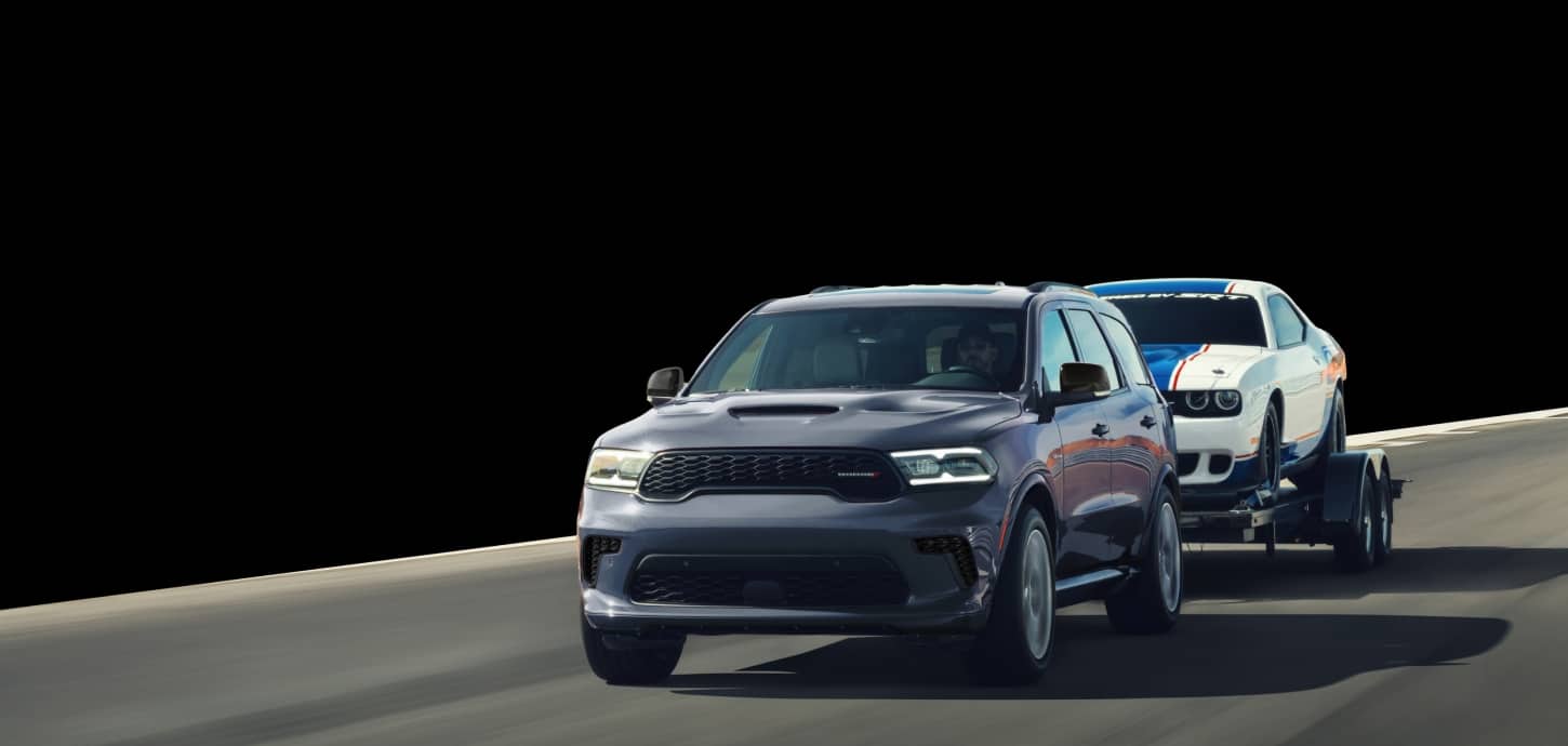 A gray 2026 Dodge Durango GT HEMI traveling down a road, towing a white Dodge Charger with a large blue racing center stripe. The background is blacked out.