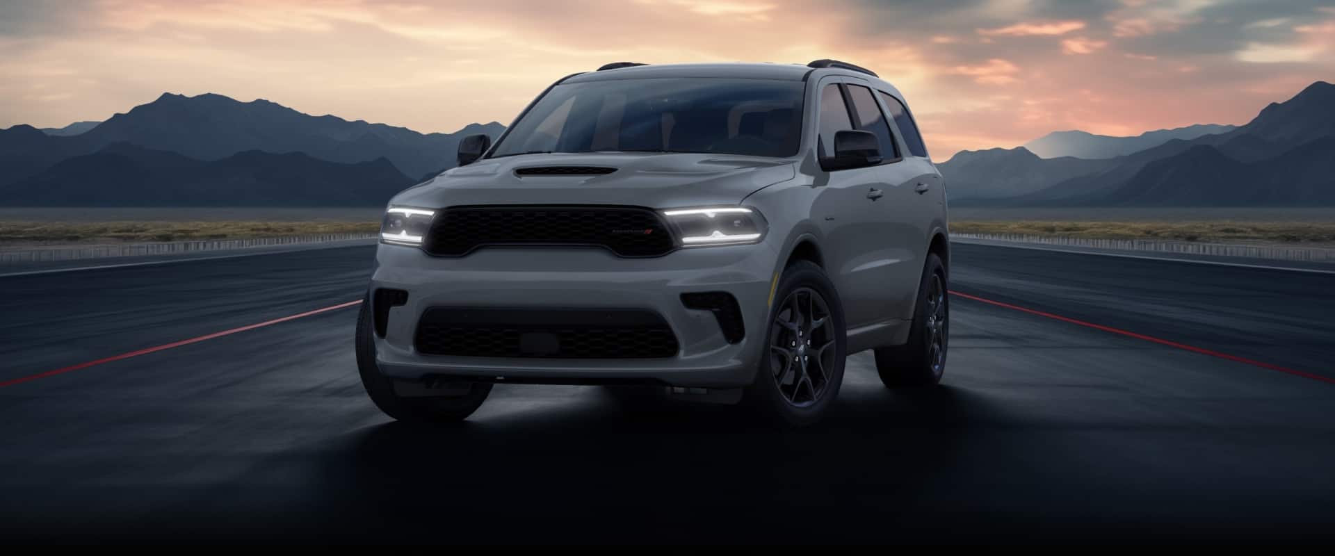 A front angle of a silver 2026 Dodge Durango GT HEMI parked on a track at sunset, surrounded by mountains.