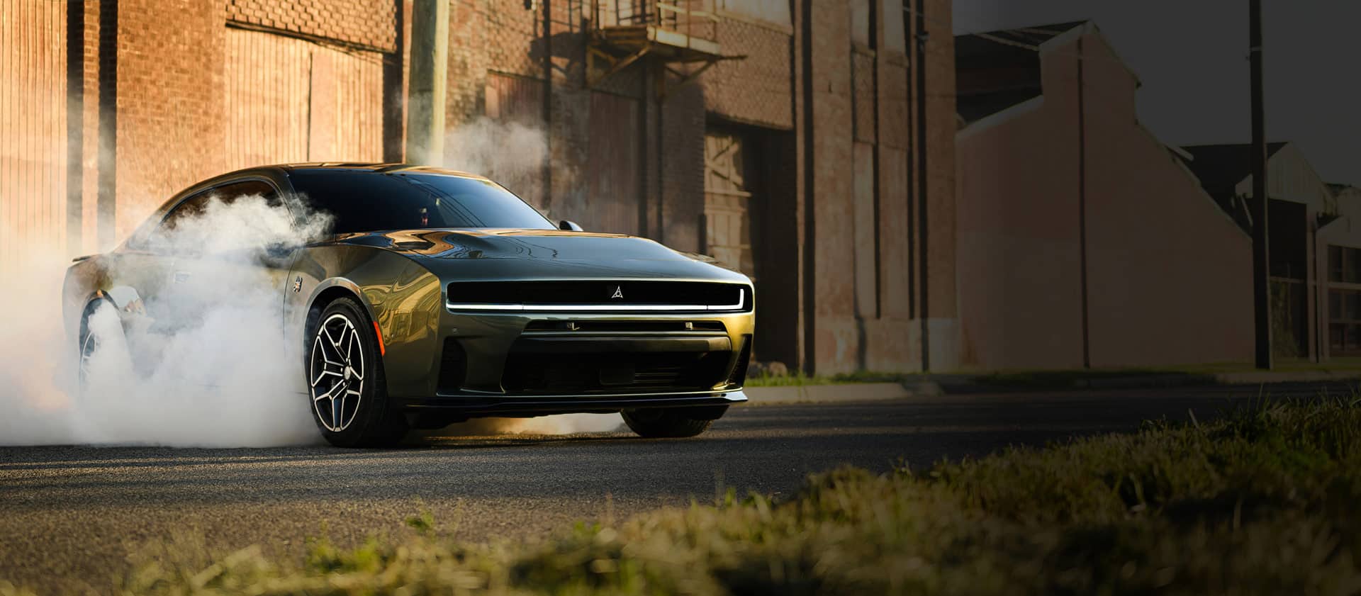 A passenger-side front angle of a black 2026 Dodge Charger Scat Pack Plus parked on blacktop with smoke billowing from its tires. In the background are industrial buildings.