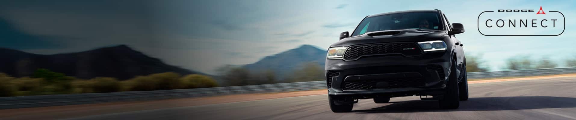 A head-on angle of a black 2026 Dodge Durango GT traveling on a curved racetrack. The Dodge Connect logo.